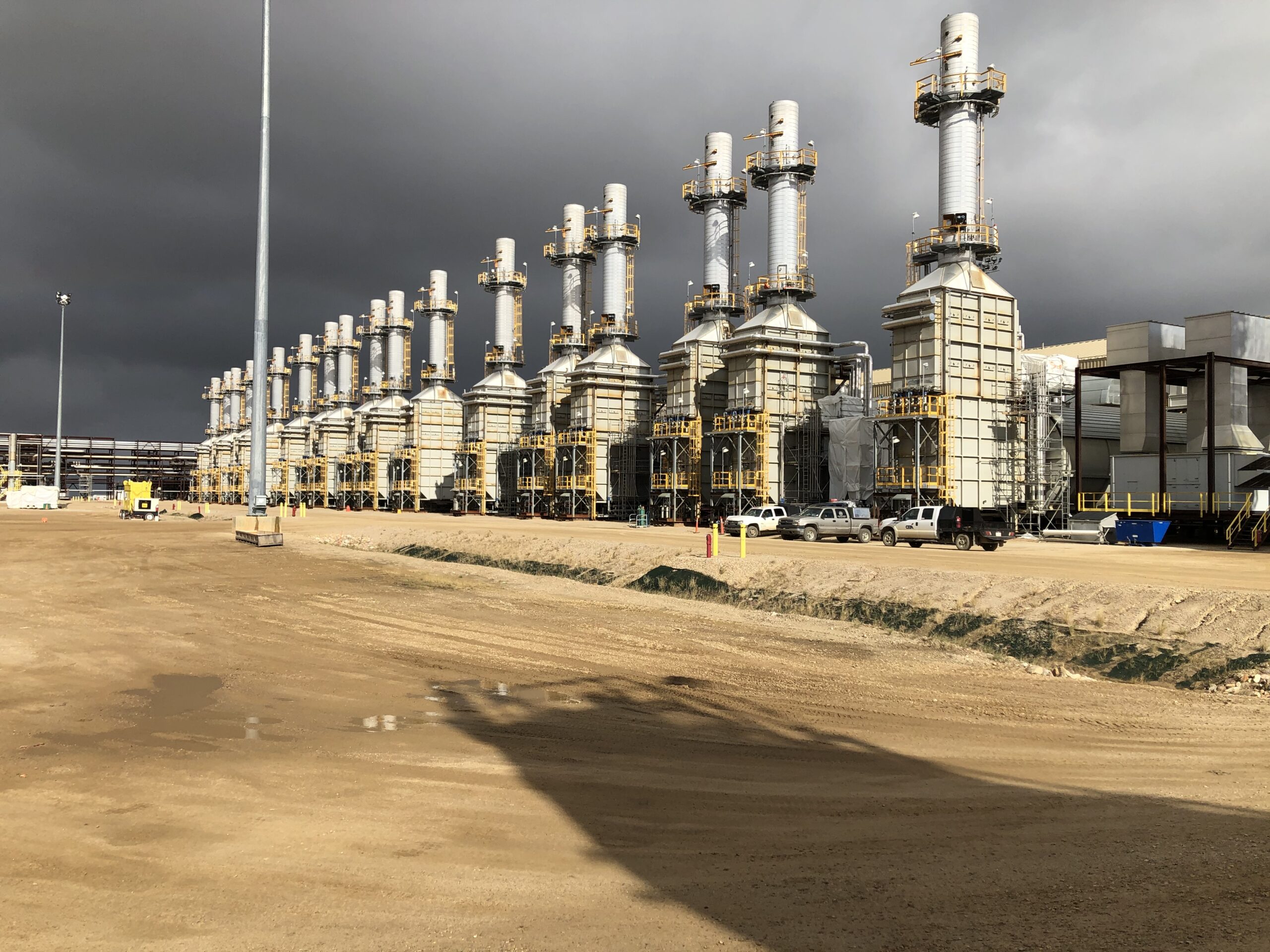 Interior view of an oil and gas facility, filled with complex machinery, pipes, control panels, and operators in safety gear overseeing operations.