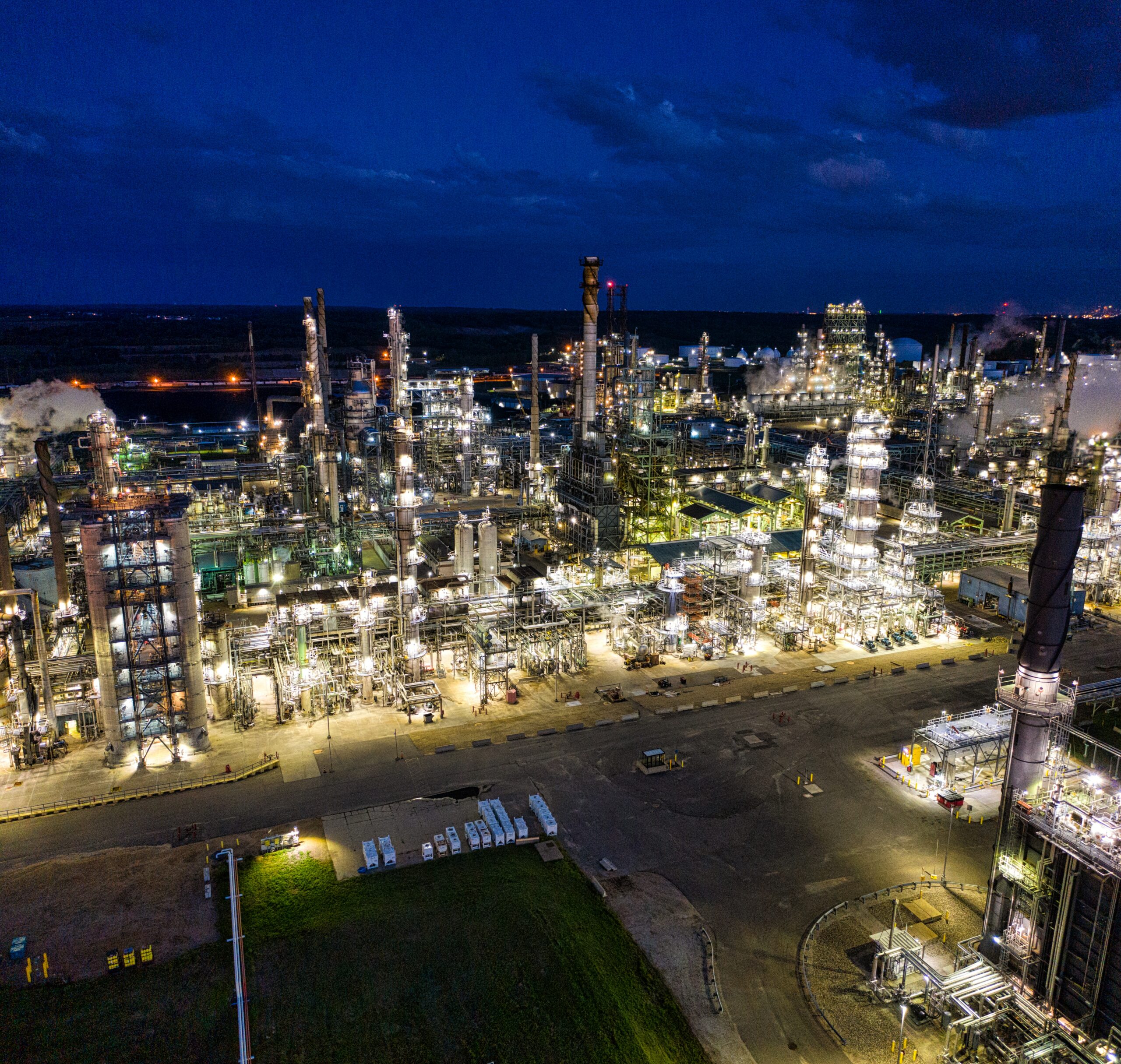 Aerial nighttime view of an oil and gas facility, brightly illuminated, with glowing lights outlining storage tanks, pipelines, and industrial structures, all amidst the surrounding darkness.