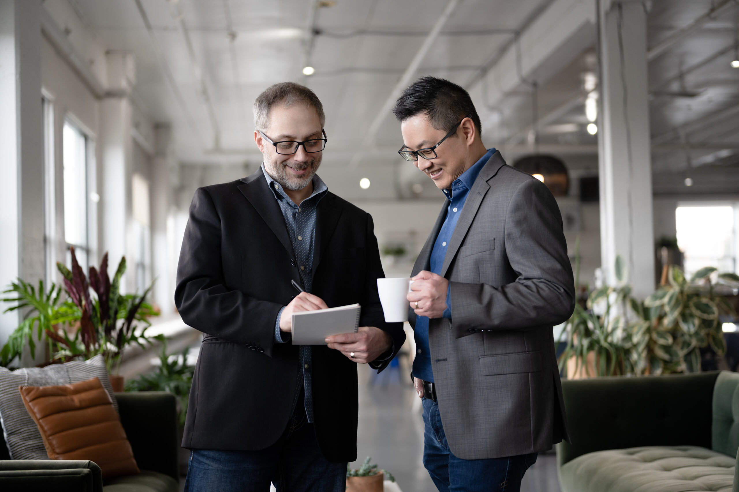 Picture displaying two SysEne colleagues. One of them is writing in a notepad smiling and showing what he's writing to his colleague. His colleague is holding a coffee cup but looks very interested in what's being said.