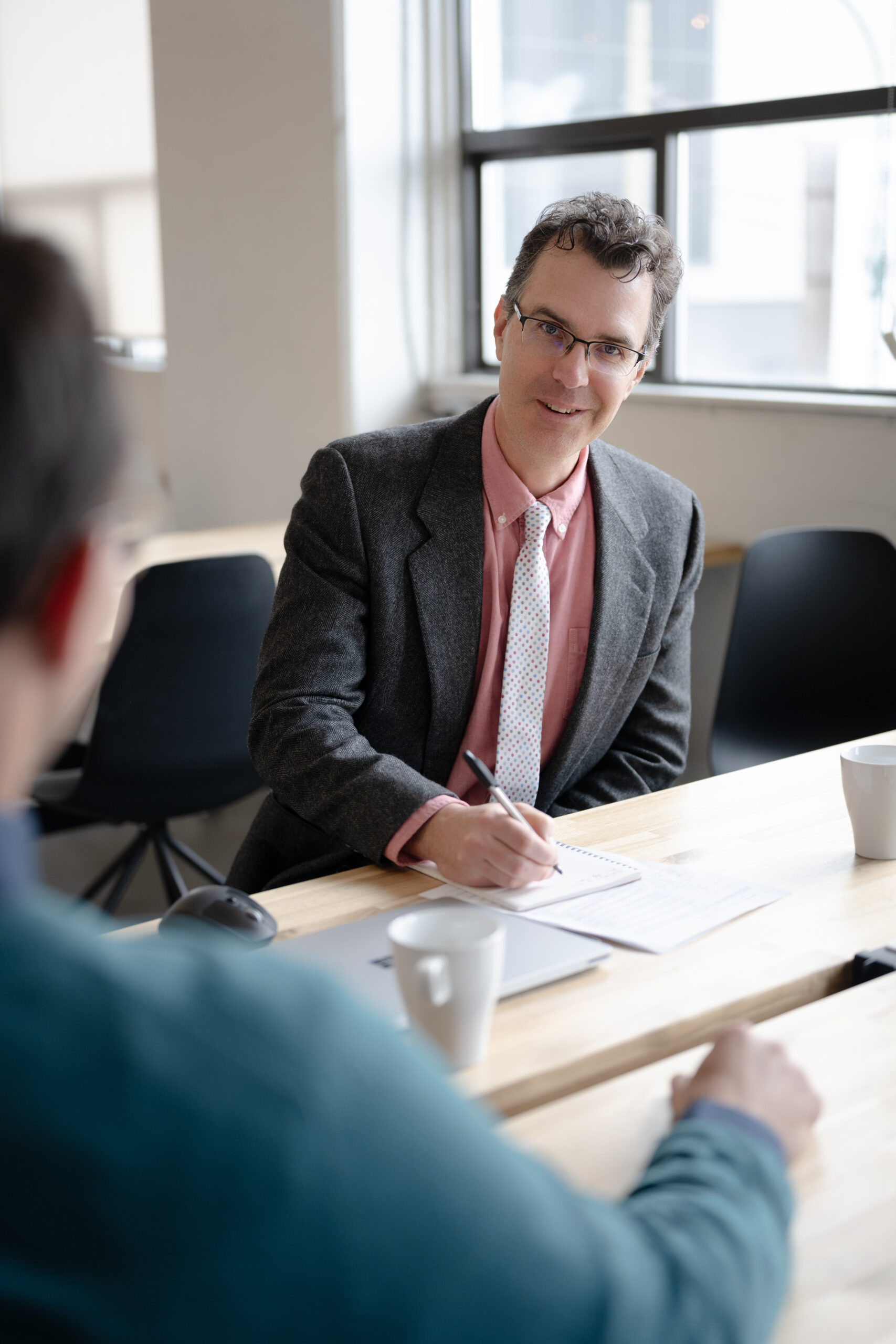 SysEne consultant attentively engaged with client during face to face meeting. SysEne consultant is poised ready to take notes and has a smile on his face as he listens.